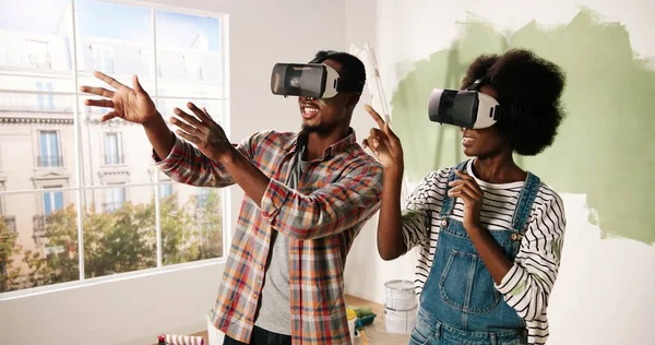Porträt eines fröhlichen jungen afroamerikanischen Ehepaares, das bei Renovierungsarbeiten mit einer VR-Brille im Zimmer steht und das erneuerte Wohnungsdesign mithilfe futuristischer Virtual-Reality-Technologie betrachtet — Stockfoto