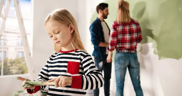 Close up portret uroczej szczęśliwej dziewczynki uśmiecha się do aparatu stojącego w pokoju z pędzlem w rękach. Mama i tata malują ścianę w zielonym kolorze na tle. Naprawa domu. Projekt wnętrza — Zdjęcie stockowe