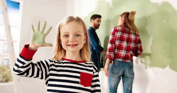 Närbild porträtt av glad liten söt flicka ler mot kameran visar handflatan i grön färg. Mamma och pappa målar vägg på bakgrund. Reparation och ombyggnad av hemmet. Förnya huset. Inredning — Stockfoto
