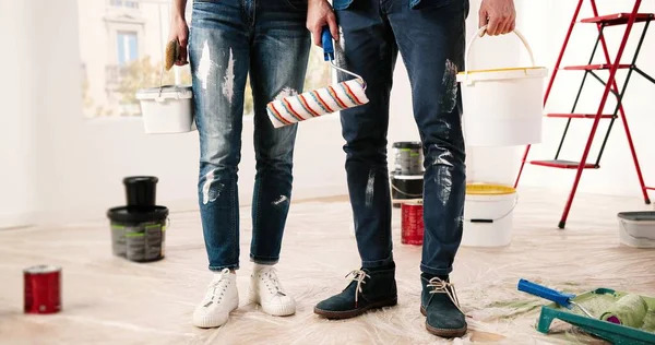 Zblízka mužské a ženské nohy stojící v bytě při opravách. Muž a žena drží barvu a kolečko štětec v místnosti před rekonstrukcí a přestavbou domu. Dekor cocnept — Stock fotografie