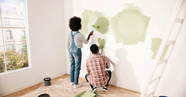 Zadní pohled na mladé Afroameričany manželé pár muž a žena pracující v pokoji renovace vlastního domu. Manželka a manžel malují stěny v zelené barvě společně pomocí válečkových štětců. koncept domácí opravy — Stock fotografie