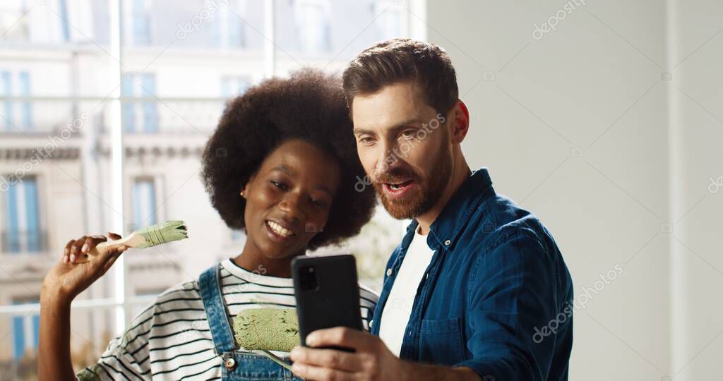 Portrait of happy mixed-races young married family couple man and woman smiling in new apartment taking selfie photos on smartphone posing with brushes in room. Home repair concept