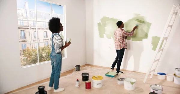 Bakre delen av unga afroamerikanska par arbetar i rummet omformar egen lägenhet. Frun pratar med maken om att måla väggar. Handyman målar vägg i grön färg med roller borste reparation koncept — Stockfoto
