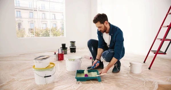 Close up of Caucasian young handsome man pouring green olive paint preparing for painting walls in house renovating and redesigning room. Handyman working on apartment redecoration repair and makeover