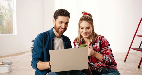 Ritratto di felice caucasico giovane coppia moglie e marito seduto sul pavimento in appartamento, parlando e scegliendo nuova casa di design navigazione online utilizzando il computer portatile. sorseggiando da bere. Riparazione casa — Foto Stock