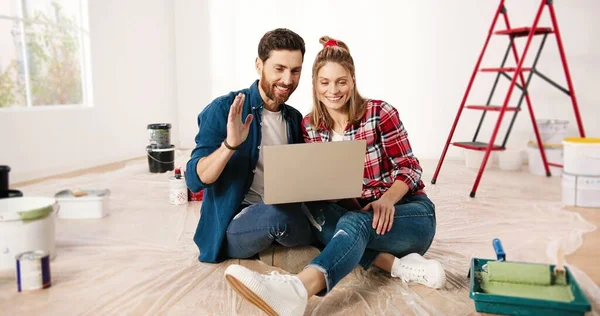 Vrolijke blanke jonge getrouwde echtpaar en echtgenoot zitten op de vloer in appartement in de buurt ladder, spreken op video online bellen met behulp van laptop computer. Huisreparatie concept. Videochatting op webcam — Stockfoto