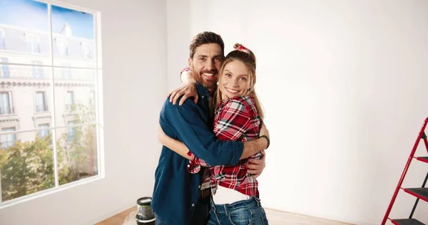 Macchina fotografica si avvicina felice giovane coppia caucasica uomo e donna che si abbracciano mentre in piedi in appartamento vuoto vicino a una grande finestra durante il processo di riparazione a casa e sorridendo alla fotocamera. Concetto d'amore. Da vicino. — Foto Stock