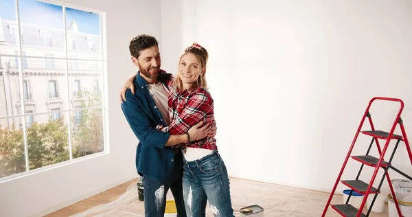 Camera nadert gelukkige jonge blanke echtpaar man en vrouw knuffelen terwijl staan in leeg appartement in de buurt van groot raam tijdens het huis reparatieproces en glimlachen naar de camera. Liefdesconcept. Sluiten. — Stockfoto
