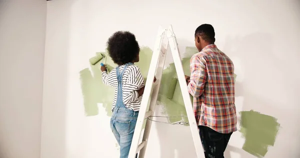 Back view of African American couple male and female redesigning and renovating room in own apartment standing on ladder painting walls in green olive color with roller brush. Home repair concept. — Stock Photo, Image