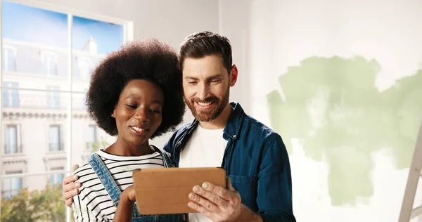Close-up portret van vrolijke multi-etnische jonge echtpaar Kaukasische man en Afro-Amerikaanse vrouw staan in de kamer typen op tablet apparaat bespreken ontwerp van nieuw huis. Huisreparatie concept — Stockfoto