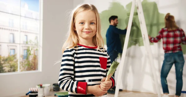 Primer plano retrato de adorable niña linda feliz sonriendo a la cámara de pie en la habitación con pincel de pintura en las manos. Mamá y papá pintando la pared en color verde sobre fondo. Reparación. Diseño de interiores — Foto de Stock