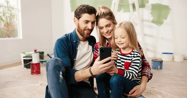 Vreugdevolle jonge zorgzame blanke ouders zitten met kleine schattige kleuter meisje op de vloer na het schilderen van muren chatten, tikken en surfen online op de smartphone, het kiezen van nieuwe kamer decor. Huisreparatie — Stockfoto