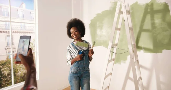 Afro-americano encantador marido tirar fotos de bela esposa feliz no quarto fazendo renovação em casa posando com pincel nas mãos. Renovação e conceito de reparação em casa. Fotografar no celular — Fotografia de Stock