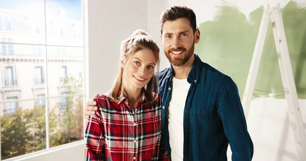 Macchina fotografica si avvicina felice giovane coppia caucasica uomo e donna che si abbracciano mentre in piedi in appartamento vuoto vicino a una grande finestra durante il processo di riparazione a casa e sorridendo alla fotocamera. Concetto d'amore. Da vicino. — Foto Stock