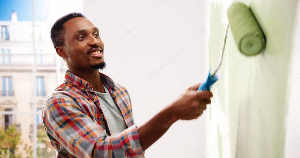 Close up portrait of happy young African American man standing in new apartment renovating room painting wall with brush roller in hand renewing home. Repair concept. Decorating and repairing
