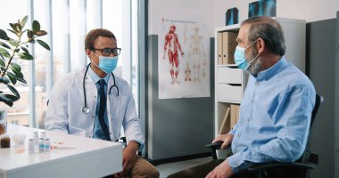 Mutlu Afrikalı genç yakışıklı erkek doktor, tıbbi maske uzmanı. Kafkasyalı yaşlı hastayla konuşuyor. Coronavirus aşısı aşısı konseptini gösteriyor ve açıklıyor.
