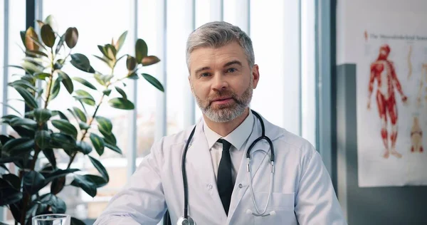 Ritratto ravvicinato di bel caucasico allegro positivo anziano medico maschio in camice medico bianco guardando la fotocamera e sorridendo in armadietto in clinica, uomo specialista sanitario, concetto di ospedale — Foto Stock