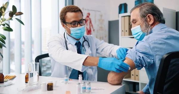 Close up af afrikansk ung smuk mand læge i medicinsk maske gør vaccine injektion til kaukasiske senior patient sidder på hospitalet kabinet lab, coronavirus vaccination koncept - Stock-foto