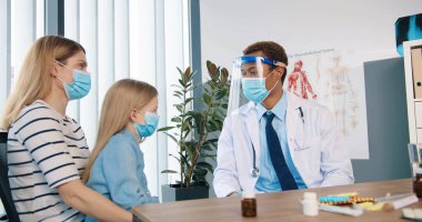 Afrikalı Amerikalı yakışıklı genç adam portresi. Koruyucu yüz kalkanı ve tıbbi maske takan doktor. Klinikte oturmuş, beyaz kadın hasta ve küçük çocukla konuşuyor.