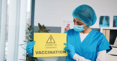 Hindu genç kadın uzman hemşire portresi. Koruyucu eldivenler ve tıbbi maske takıyor. Elinde sarı kağıt kart var.