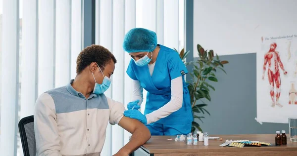 Kvindelig ung professionel sundhedspleje assistent sygeplejerske i beskyttende uniform forbereder injektion og injicere covid-19 vaccine til smuk mandlig patient i medicinsk maske sidder på hospitalet lab room - Stock-foto