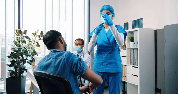 Bageste af Hindu unge kvindelige sygeplejerske specialist i uniform gør coronavirus vaccine injektion til mandlige patient sidder i kabinet på hospitalet, mens lægen arbejder bag, covid vaccination koncept - Stock-foto