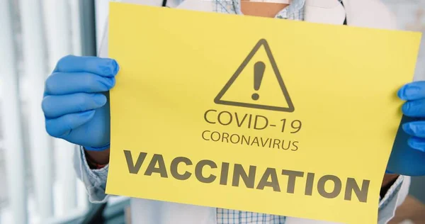Close up skud af person hænder i beskyttelseshandsker holder papir kort med meddelelse om coronavirus vaccination kampagne, covid-19 vaccine, opmærksomhed, sundhedsmeddelelse - Stock-foto