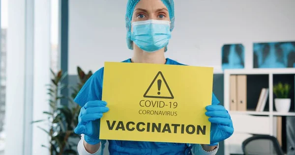 Close up af kaukasiske smukke kvindelige specialist læge i beskyttelseshandsker og medicinsk maske holder i hænder gult papir kort, der viser meddelelse om covid-19 vaccination, coronavirus pandemi - Stock-foto