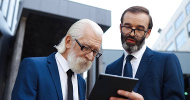 Kafkasyalı iki işadamı iş merkezinin önünde duruyor ve tablet aygıt kullanıyorlar. Orta yaşlı bir adam eski meslektaşına bilgisayarında bir şey gösteriyor ve iş konuşuyor..