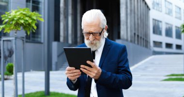 Beyaz saçlı, hava kapsüllü, gözlüklü, açık havada duran ve video kaset aracılığıyla tablet cihazıyla konuşan yaşlı bir adam. Büyükbaba iş adamı caddede bir aletin üzerinde görüntülü sohbet yapıyor. iş kavramı.