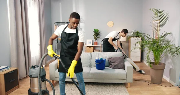 Close up of African American young female and male professional cleaners  vacuuming floor with vacuum in modern room, cleaning service, small  business, working in apron, clean house concept Stock Photo