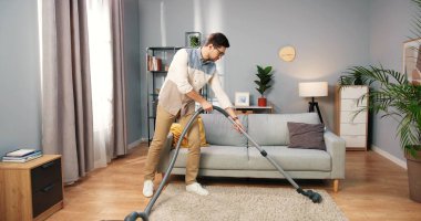 Beyaz tenli yakışıklı ciddi bir genç adam halı süpürüyor modern apartman temizliyor, temizlik yapıyor, temizlikçi, ev işlerini yapıyor ve oturma odasını elektrik süpürgesiyle temizliyor.