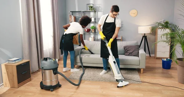 Cheerful young multi-ethnic male and female professional workers working in aprons vacuuming floor and sofa in clients home, cleaning service industry concept, small business, job
