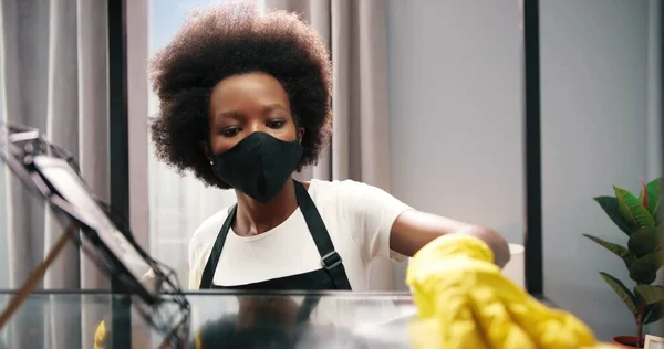 Close up of pretty young African American female worker in mask, apron and gloves working in living room and cleaning home, cleaning service concept, disinfecting house microbes, apartment hygiene
