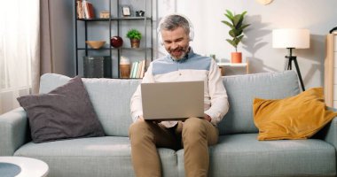 Neşeli, beyaz, orta yaşlı, sakallı bir adam rahat bir odada, kulaklıkla oturmuş dizüstü bilgisayar başında komedi filmi ya da videolar izliyor ve gülüyor, pozitif duygular, boş zaman kavramı.