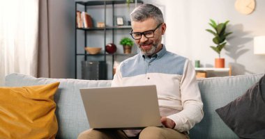 Gözlüklü, beyaz, neşeli, gözlüklü, dizüstü bilgisayarlı, mutlu bir şekilde internette yazarken rahat, modern bir oturma odasında tek başına, evden uzak bir yerde, boş bir konseptte oturuyor.