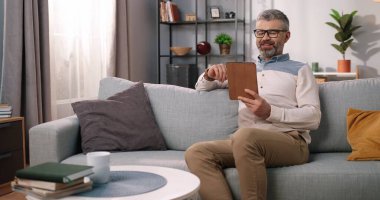 Beyaz sakallı, orta yaşlı, gözlüklü, mutlu, olgun bir adam. Kanepede oturmuş, pozitif ruh halinde. Tablet cihazına tıklıyor. Oturma odasında, boş zamanlarında, ev kavramında videolar izliyor.