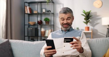 Neşeli, orta yaşlı, sakallı, rahat bir odada koltukta oturan, akıllı telefonuyla internetten kredi kartı alan ve EVET jesti, e-ticaret, alışveriş yapan beyaz adamın yakın plan fotoğraflarını çek.