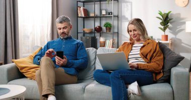 Dijital aletlerle evdeki kanepede dinlenen mutlu evli çift. Orta yaşlı, beyaz bir adam akıllı telefondan mesaj yazarken güzel karısı internette bilgisayarını karıştırıyor. Teknoloji kavramı.