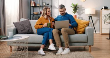 Beyaz, orta yaşlı, evli bir çift evlerinde kanepede oturmuş internetten alışveriş yapıyorlar. Laptop bilgisayarında kredi kartı ile ödeme yapıyorlar. Konforlu bir odada, e-ticarette, online alışveriş kavramında EVET hareketi yapıyorlar.