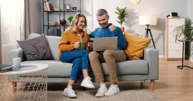 Beyaz, orta yaşlı, evli bir çift evlerinde kanepede oturmuş internetten alışveriş yapıyorlar. Laptop bilgisayarında kredi kartı ile ödeme yapıyorlar. Konforlu bir odada, e-ticarette, online alışveriş kavramında EVET hareketi yapıyorlar.