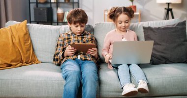 Kafkasyalı şirin çocuklar kanepede oturmuş dijital aletlerle internette geziniyorlar. Küçük tatlı kız dizüstü bilgisayarda sörf yaparken çocuk tablet cihazıyla oyun oynuyor, aile kavramı.