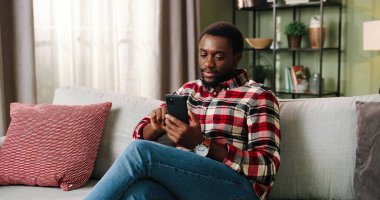 Neşeli genç Afro-Amerikan milenyum adamı elinde akıllı telefonla oturma odasındaki koltukta oturan mobil uygulamayı kullanarak sosyal medya hikayeleri videosu izliyor. Evde dinlenirken cep telefonu kullanan mutlu adam.