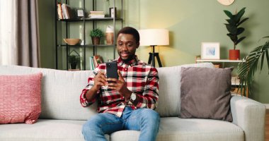 Yakışıklı, Afro-Amerikan, mutlu, genç bir erkek, evdeki kanepede oturuyor ve internetteki konuşmasında akıllı telefondan olumlu bir şekilde el sallıyor. Çevrimiçi konuşma konsepti