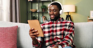 Beyaz kulaklıklı genç Afrikalı bir adam evdeki koltukta oturup arkadaşlarıyla tablet kullanarak sohbet ediyor..