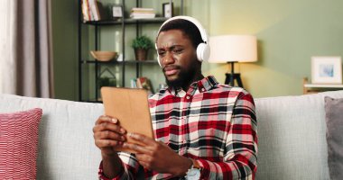 Beyaz kulaklıklarla müzik dinleyen yakışıklı, Afro-Amerikan genç bir adamın portresi rahat evdeki koltukta tek başına oturup tablet aygıt kullanarak en sevdiği şarkıları dinliyor. Ev kavramı