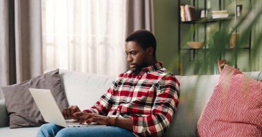 Afro-Amerikalı yakışıklı, yakışıklı, ciddi bir adam oturma odasında oturmuş bilgisayarında daktilo yazıyor dizüstü bilgisayarında çalışıyor, yoğun bir yüzü var. Serbest çalışma, evde öğrenim, eğitim kavramı