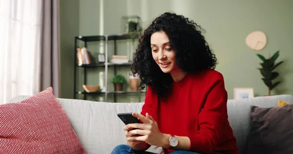 Портрет расслабленной кавказской миллениальной красавицы, которая держит смартфон и смотрит видео истории из социальных сетей, сидя на диване в гостиной. Счастливая юная леди с мобильными приложениями отдыхает дома — стоковое фото