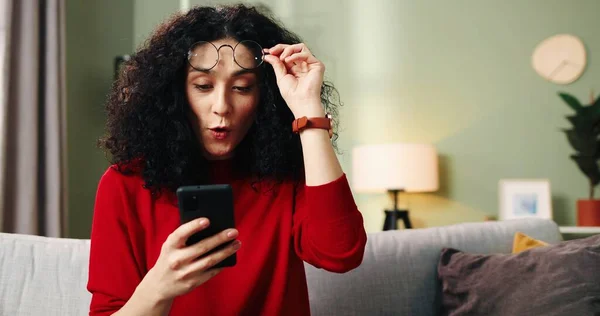 Молодая красивая женщина сидит дома и читает сообщение на смартфоне. Концепция получения хороших новостей, неожиданный сюрприз. — стоковое фото
