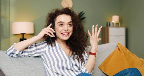 Портрет кавказской молодой позитивной эмоциональной симпатичной молодой девушки с вьющимися волосами, сидящей на диване в уютной квартире и болтающей по мобильному телефону, разговаривающей по телефону на смартфоне в хорошем настроении — стоковое фото
